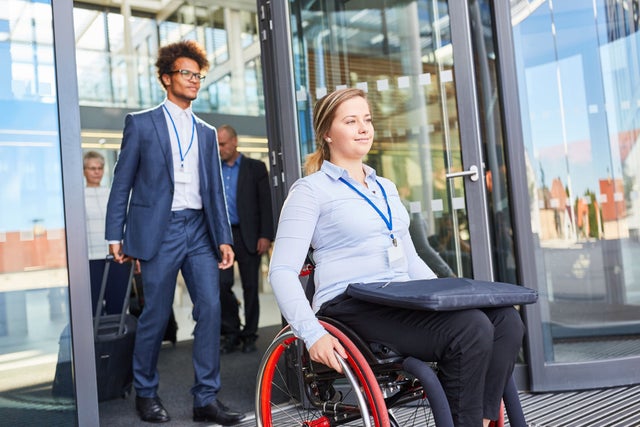 A woman in a wheelchair exiting the door of a building with men walking behind her.