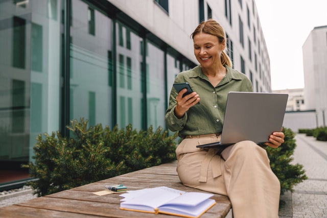 A woman sitting on a bench using both a laptop and a smartphone