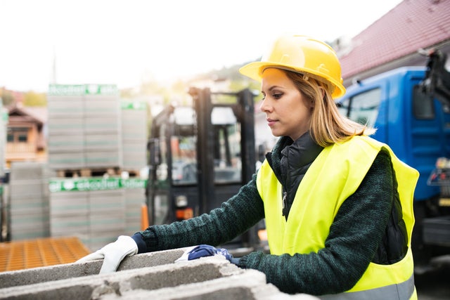 A construction worker in a yellow helmet and a vest placing tiles with crane in the background.