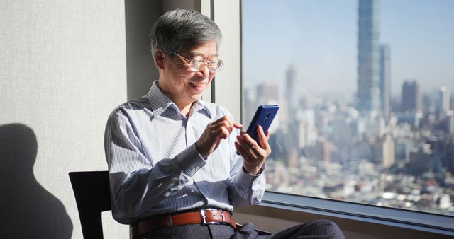 An older man sitting in front of a window looking at a phone in his hands.