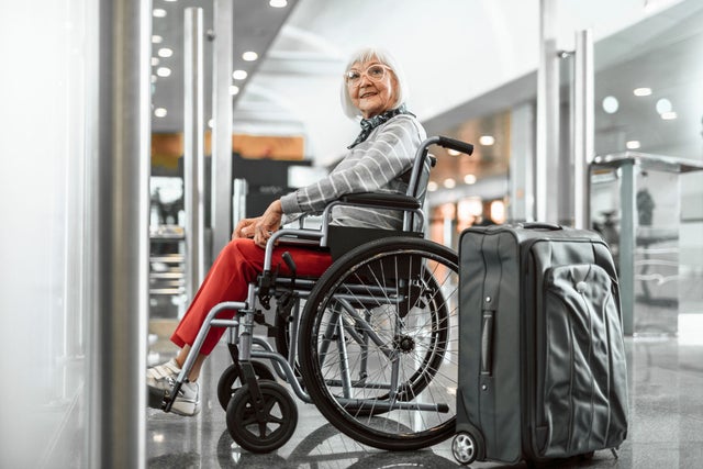 Full length portrait of mature female with laptop and suitcase sitting in wheelchair at airport and waiting for trip