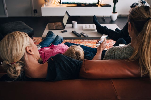 From behind a family sitting on a couch with a computer on the coffee table and one person holding a phone.