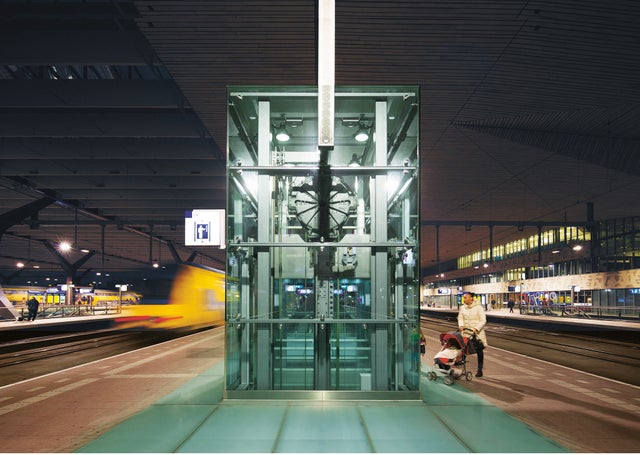 A woman waiting for an elevator outside on a train platform, elevator is in the middle and on the left side the train is leaving the station.