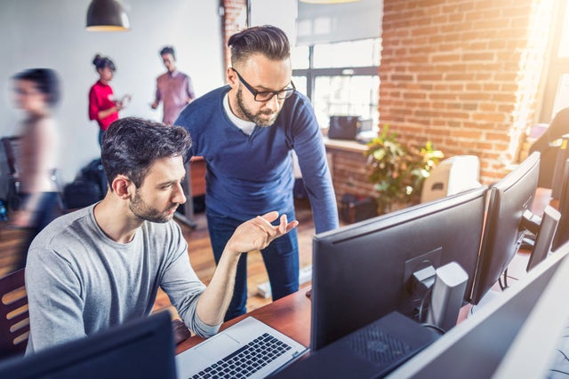 Two people looking at a computer screen and discussing, other people flow in the background.