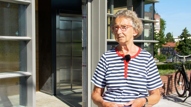 An older woman wearing a striped shirt is being interviewed with elevator in the background.