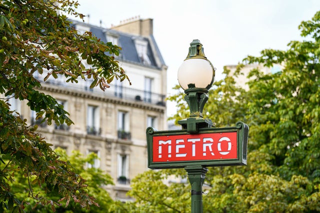 Traditional and iconic Paris metro (metropolitain, subway) sign with trees and a parisian Haussmann apartment block in the background; Shutterstock ID 2025845057